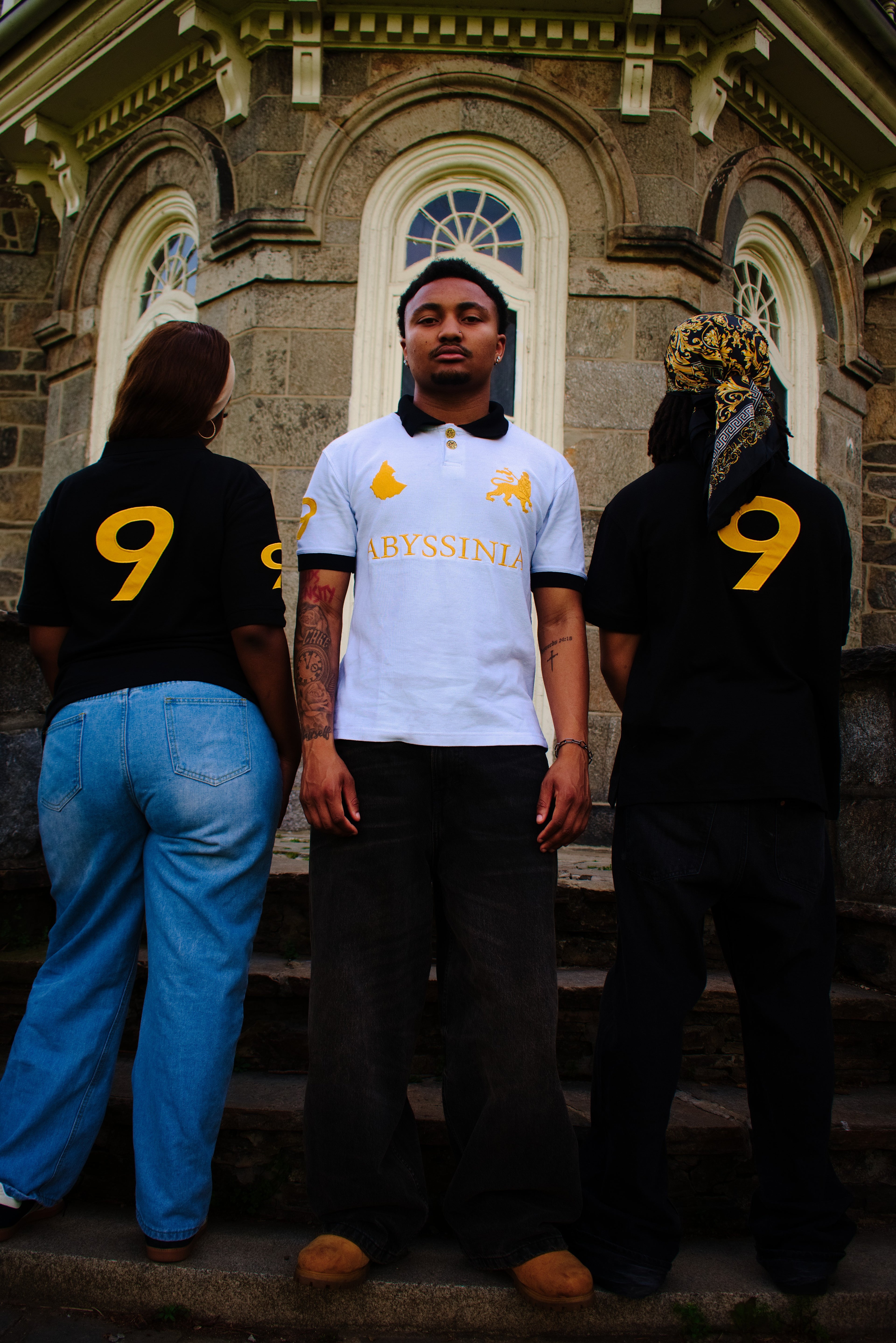 Three people standing on steps in front of a stone building, two wearing black shirts with yellow numbers and one in a white shirt.